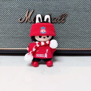 K&ouml;nnte beinhalten: Eine kleine Pl&uuml;schfigur in einem roten Liverpool FC-Outfit, einschlie&szlig;lich Hut, Schal und Pullover. Die Figur hat schwarzes Fell und wei&szlig;e Ohren. Der Schal hat ein rot-wei&szlig;es Muster. Der Pullover hat das Standard Chartered-Logo.