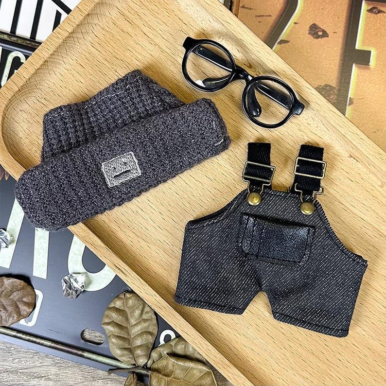 K&ouml;nnte beinhalten: Ein Miniatur-Outfit-Set mit einer dunkelgrauen Strickm&uuml;tze mit quadratischem Aufn&auml;her, einer schwarzen Jeans-Latzhose mit verstellbaren Tr&auml;gern und einer kleinen runden schwarzen Brille. Die Artikel sind auf einem Holztablett angeordnet.