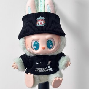 K&ouml;nnte beinhalten: Ein Pl&uuml;schtier-Hase mit schwarzer Strickm&uuml;tze mit Liverpool-Wappen und einem schwarzen Trikot mit dem Standard Chartered-Logo. Der Hase hat blaue Augen, gestickte Details und mintgr&uuml;ne Akzente.