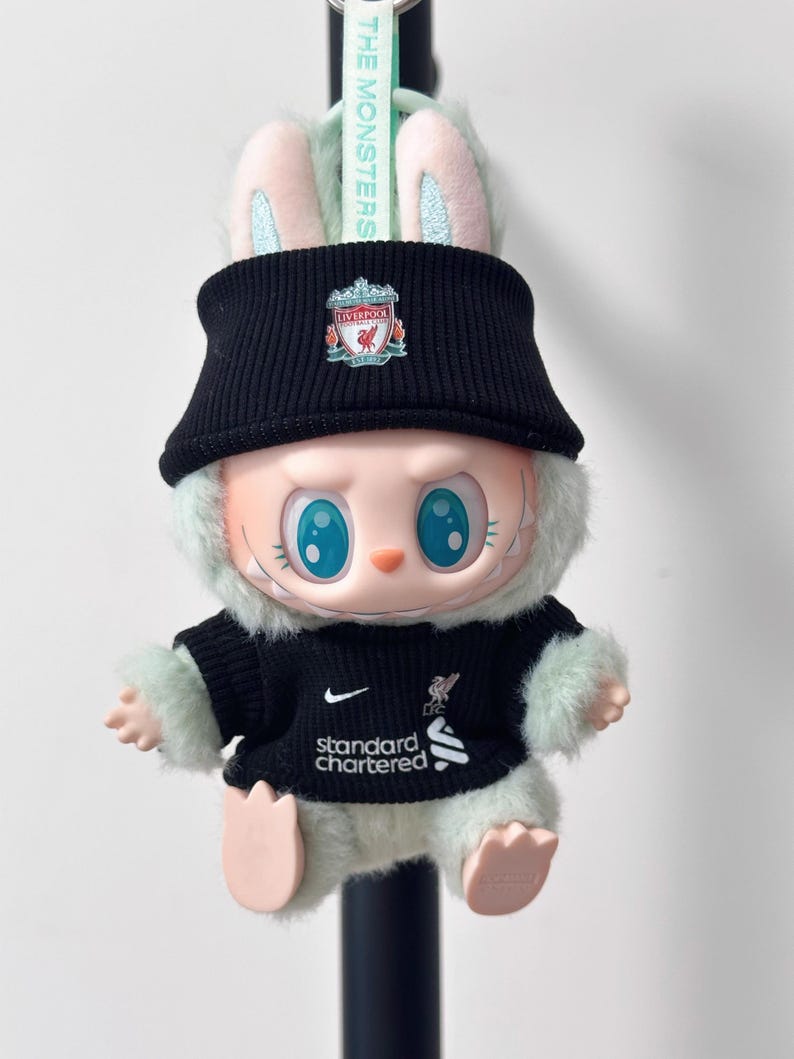 K&ouml;nnte beinhalten: Ein Pl&uuml;schtier-Hase mit schwarzer Strickm&uuml;tze mit Liverpool-Wappen und einem schwarzen Trikot mit dem Standard Chartered-Logo. Der Hase hat blaue Augen, gestickte Details und mintgr&uuml;ne Akzente.