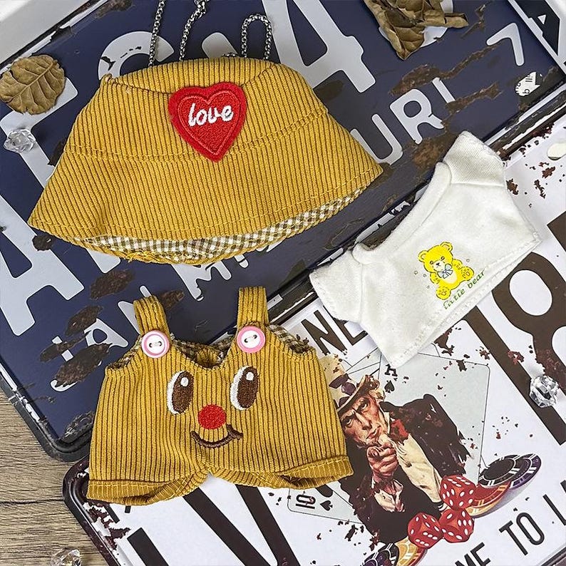 K&ouml;nnte beinhalten: Ein Set Puppenkleidung, darunter ein senfgelber Cordhut mit einem roten Herz mit der Aufschrift "love", eine passende Latzhose mit einem l&auml;chelnden Gesicht und ein wei&szlig;es T-Shirt mit einer gelben B&auml;r Grafik. Die Artikel werden auf einer Vintage-Metalloberfl&auml;che pr&auml;sentiert.