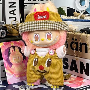 K&ouml;nnte beinhalten: Eine Pl&uuml;schfigur mit gelbem Cordhut und Latzhose. Der Hut hat einen herzf&ouml;rmigen Aufn&auml;her mit dem Wort "love". Die Figur hat rosa Fell und gro&szlig;e Augen. Eine Karte mit einem Cartoon-Hasen ist im Hintergrund zu sehen.