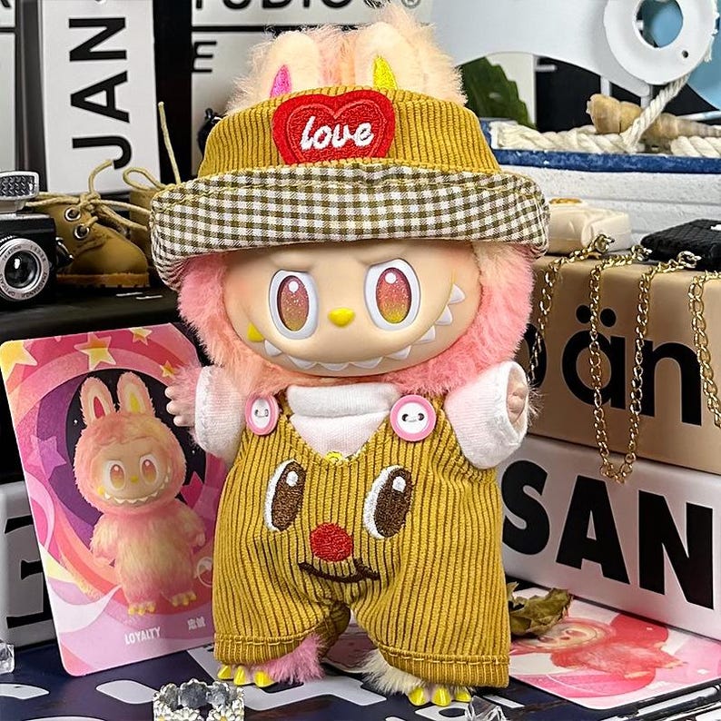 K&ouml;nnte beinhalten: Eine Pl&uuml;schfigur mit gelbem Cordhut und Latzhose. Der Hut hat einen herzf&ouml;rmigen Aufn&auml;her mit dem Wort "love". Die Figur hat rosa Fell und gro&szlig;e Augen. Eine Karte mit einem Cartoon-Hasen ist im Hintergrund zu sehen.