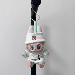K&ouml;nnte beinhalten: Eine Pl&uuml;sch-Hasenpuppe mit wei&szlig;em Fischerhut und wei&szlig;em Pullover mit Teamlogo und dem Text "Standard Chartered". Die Puppe hat blaue Augen, rosa Ohren und hellgr&uuml;ne Fellbes&auml;tze. Ein Etikett lautet "THE MONSTERS".
