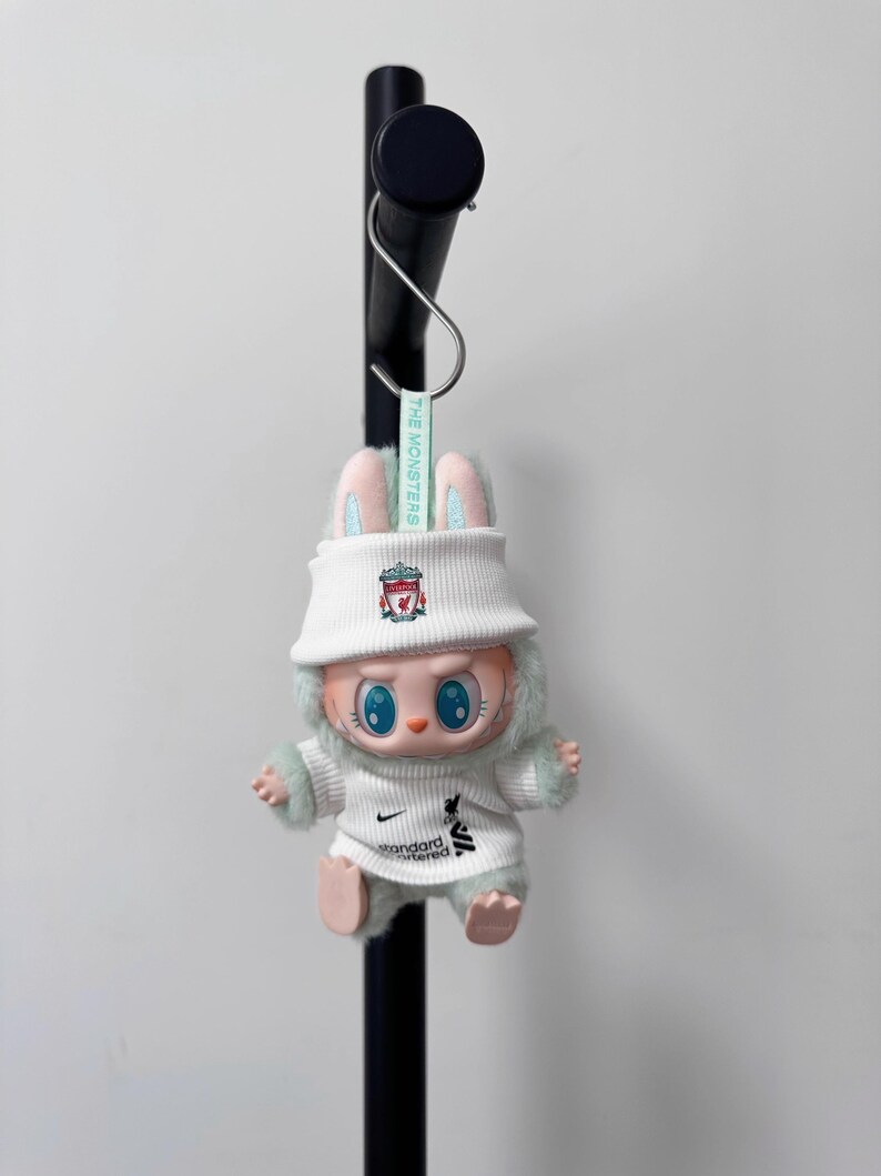 K&ouml;nnte beinhalten: Eine Pl&uuml;sch-Hasenpuppe mit wei&szlig;em Fischerhut und wei&szlig;em Pullover mit Teamlogo und dem Text "Standard Chartered". Die Puppe hat blaue Augen, rosa Ohren und hellgr&uuml;ne Fellbes&auml;tze. Ein Etikett lautet "THE MONSTERS".