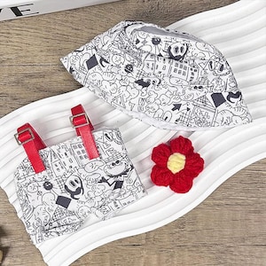 K&ouml;nnte beinhalten: Ein wei&szlig;er Fischerhut und ein passendes Romper-Set mit einem schwarz-wei&szlig;en Cartoon-Design. Der Romper hat rote Tr&auml;ger und Schnallen. Eine rote geh&auml;kelte Blume mit gelbem Zentrum ist ebenfalls vorhanden.