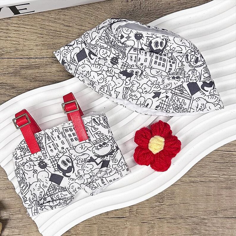 K&ouml;nnte beinhalten: Ein wei&szlig;er Fischerhut und ein passendes Romper-Set mit einem schwarz-wei&szlig;en Cartoon-Design. Der Romper hat rote Tr&auml;ger und Schnallen. Eine rote geh&auml;kelte Blume mit gelbem Zentrum ist ebenfalls vorhanden.