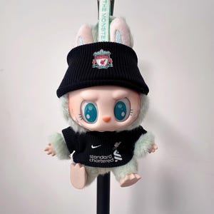 K&ouml;nnte beinhalten: Eine Pl&uuml;schpuppe mit schwarzem Liverpool FC-Hut und -Trikot. Die Puppe hat gro&szlig;e blaue Augen, rosa Ohren und einen flauschigen gr&uuml;nen K&ouml;rper. Das Trikot hat das Standard Chartered-Logo. Ein gr&uuml;nes Band mit dem Text "THE MONSTER" ist befestigt.