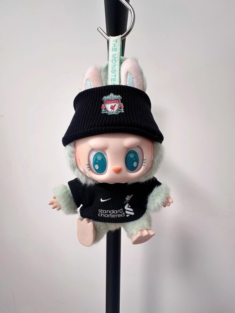 K&ouml;nnte beinhalten: Eine Pl&uuml;schpuppe mit schwarzem Liverpool FC-Hut und -Trikot. Die Puppe hat gro&szlig;e blaue Augen, rosa Ohren und einen flauschigen gr&uuml;nen K&ouml;rper. Das Trikot hat das Standard Chartered-Logo. Ein gr&uuml;nes Band mit dem Text "THE MONSTER" ist befestigt.