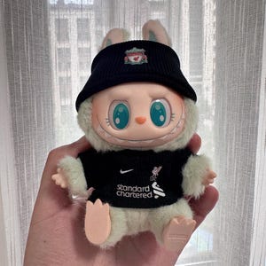 K&ouml;nnte beinhalten: Ein Pl&uuml;schtier-Hase mit schwarzer Kappe und schwarzem Trikot mit dem Nike-Logo und dem Text "standard chartered". Der Hase hat gro&szlig;e blaue Augen und wird in einer Hand gehalten.