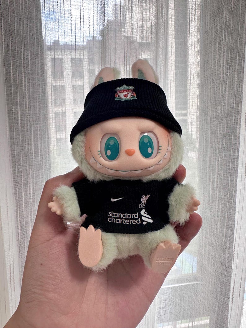 K&ouml;nnte beinhalten: Ein Pl&uuml;schtier-Hase mit schwarzer Kappe und schwarzem Trikot mit dem Nike-Logo und dem Text "standard chartered". Der Hase hat gro&szlig;e blaue Augen und wird in einer Hand gehalten.