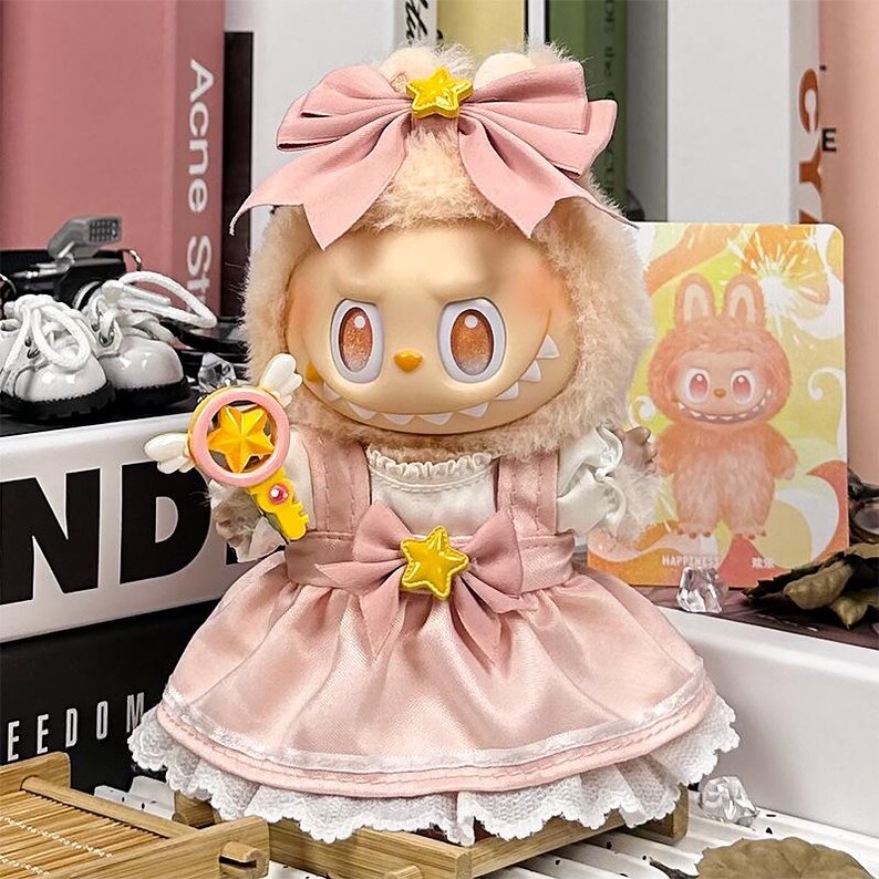 K&ouml;nnte beinhalten: Eine Sammlerpuppe mit einem skurrilen Design. Sie hat ein hellrosa Kleid mit wei&szlig;er Spitzenborte, eine gro&szlig;e rosa Schleife und ein sternf&ouml;rmiges Accessoire. Die Puppe h&auml;lt einen gelben sternf&ouml;rmigen Zauberstab mit Fl&uuml;geln.