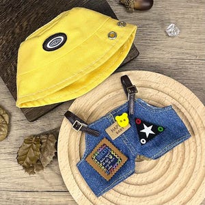 K&ouml;nnte beinhalten: Ein gelber Fischerhut mit einem schwarz-wei&szlig;en Emblem und eine Jeans-Latzhose mit braunen Tr&auml;gern. Die Latzhose hat einen B&auml;renknopf und ein Etikett mit der Aufschrift "HAPPY BABY".