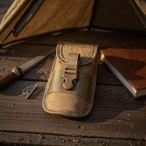 Pode incluir: Uma bolsa utilitária bege com fecho de fivela, uma faca pequena, um anel e um caderno com capa de couro estão dispostos sobre uma superfície de madeira. A bolsa tem uma aba e uma alça com fivela de plástico. A faca tem um cabo de madeira.