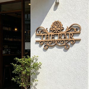 以下が含まれることがあります： 「THE BAR」の文字とバラのデザインが施された木製の看板。白いテクスチャの壁に取り付けられています。入り口の前には鉢植えの植物があり、開いたドアからバーの内部が見えます。