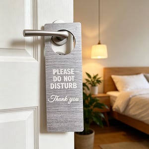 May include: A gray door hanger with the text "PLEASE DO NOT DISTURB Thank you" in white lettering. The hanger is hanging on a white door with a silver handle. The background shows a bedroom with a bed and a lamp.
