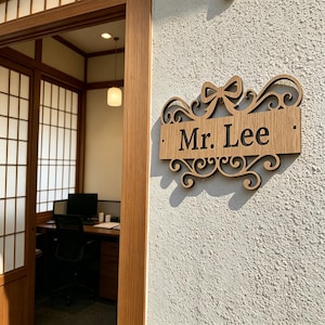 以下が含まれることがあります： 「Mr. Lee」の文字が装飾的なフォントで書かれた木製の看板。リボンとスクロールワークのデザインで、質感のある白い壁に取り付けられています。これは、家やオフィス用の看板である可能性があります。