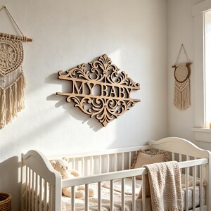 以下が含まれることがあります： 白い壁に取り付けられた、装飾的なフォントで「MY BABY」と書かれた木製の看板。 看板は、華やかなスクロールワークのデザインです。 テディベアと毛布のある白いベビーベッドが前景にあります。 マクラメの壁掛けも表示されます。