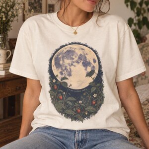 May include: An off-white short-sleeve t-shirt featuring a large circular graphic of a full moon above a strawberry patch with green leaves and red berries. The text "JUNE AFTER DARK" is printed in small white letters below the moon.
