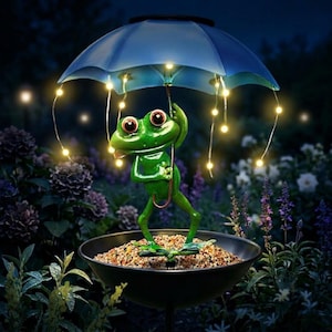 May include: A whimsical green frog statue holding a blue umbrella with string lights. The frog stands in a black birdbath filled with pebbles, set against a backdrop of flowers and foliage. A charming garden decoration.