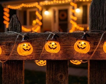 May include: A string of orange pumpkin-shaped LED lights, each wearing a small black witch hat. The pumpkins feature carved jack-o'-lantern faces that glow from within, connected by a thin wire and draped across a weathered wooden fence.