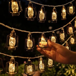 May include: String of miniature lantern lights with warm white illumination. Each lantern has a clear glass enclosure and a dark metal frame. The lights are strung on a silver wire, creating a cozy and decorative ambiance, perfect for outdoor or indoor use.