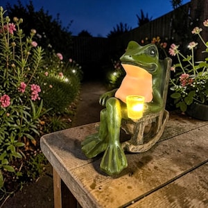 May include: A green frog statue in a rocking chair, holding a lit candle. The frog has a light-up belly and is sitting on a wooden bench. The background features a garden with pink flowers and string lights.