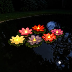 Puede incluir: Luces flotantes LED con forma de flor de loto en varios colores, incluyendo rosa, naranja, amarillo y morado, sobre hojas de nenúfar. Las luces están encendidas, creando un brillo suave en la superficie del agua. La escena es nocturna.