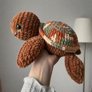 May include: A plush turtle toy crafted from brown chenille yarn with a textured finish. The shell features a variegated pattern of brown, cream, and sage green with a spiral stitch detail. It has a large green eye and a small brown tag labeled PrimeBlyss.