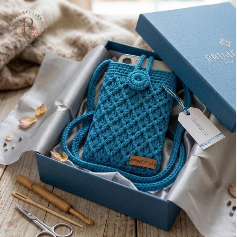 "Teal crochet phone pouch with crossbody strap and button closure, displayed in a gift box with crochet tools