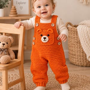 May include: A bright orange crochet overall featuring a bear face applique on the chest pocket. The garment has shoulder straps secured with light-colored buttons, matching buttons on the sides, and a textured knit pattern throughout the legs.