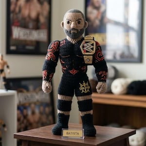 May include: A crocheted doll of a wrestler with a beard, wearing black trunks with the word EVOLUTION and a gold championship belt. The doll features red tribal-style arm tattoos, black knee pads, and black boots. A small sign in front reads BATISTA.