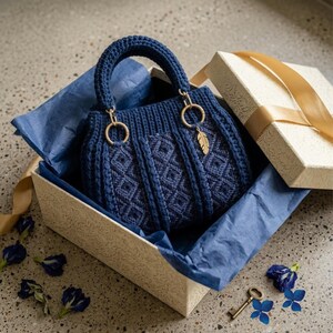 May include: A dark blue crocheted handbag with a textured diamond pattern, a rounded handle, and a gold-toned feather charm attached to a circular ring. The bag is nestled inside a cream-colored gift box lined with dark blue tissue paper.