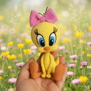 May include: A yellow crocheted bird figurine with a large head, long eyelashes, blue and black eyes, and an orange beak. The bird features a pink crocheted bow on top of its head and orange feet, crafted with a textured yarn stitch pattern.