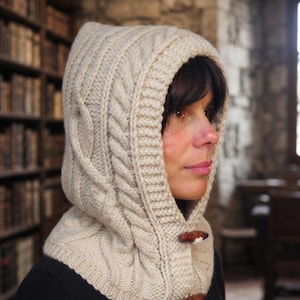 May include: A cream-colored knitted hood with a thick cable knit pattern and a ribbed border. The hood features two dark brown wooden toggle buttons fastened vertically along the front opening, providing a secure closure for the neck and head.