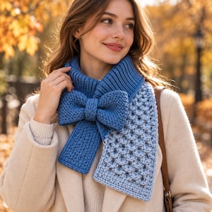 Könnte beinhalten: Ein blauer Strickschal mit einer großen, auffälligen Schleife am Hals. Der Schal zeigt eine Kombination aus Rippen- und Strukturstrickmustern in einem mittleren Blauton und wird über einem hellbeigen Mantel mit weicher Oberfläche getragen.