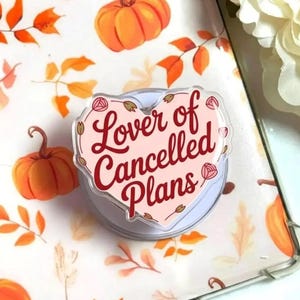 Puede incluir: Una taza de cerámica blanca con un asa negra y el texto "But First, Coffee" en negrita negra. La taza se encuentra sobre un fondo borroso, sugiriendo un ambiente acogedor.