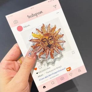 May include: A decorative sun-shaped phone grip with a smiling face, in shades of yellow, orange, and brown. The grip is on a clear base, and the image is set against a pink Instagram-themed background. The text on the background says "Instagram".