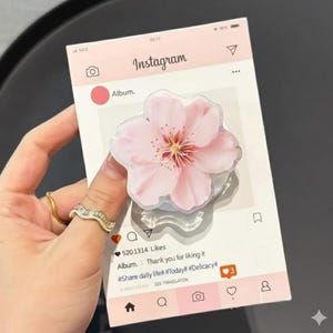May include: A pink and white Instagram-themed phone grip featuring a cherry blossom design. The grip is held by a hand, and the Instagram graphic includes the word "Instagram", a heart, and the text "Album. Thank you for liking it".