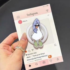 May include: A clear phone grip featuring a cartoon duck wearing sunglasses and a blue towel, with green slippers. The grip is on a pink Instagram-themed background with text that reads "Album. Thank you for liking it #Share daily life# #Today# #Delicacy#".