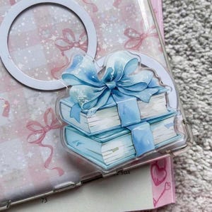 Puede incluir: Una funda transparente para teléfono con una pegatina decorativa que muestra dos libros azul claro atados con una cinta y un lazo azules. El fondo presenta un diseño rosa y blanco con lazos y un círculo blanco.