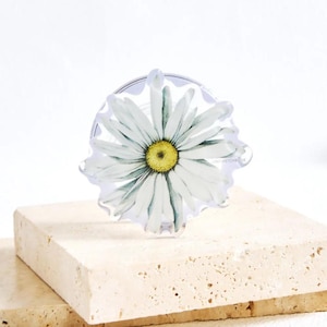 Puede incluir: Soporte de teléfono acrílico transparente con una margarita blanca y centro amarillo. El diseño floral tiene detalles verdes. El soporte está sobre una superficie de piedra beige claro.