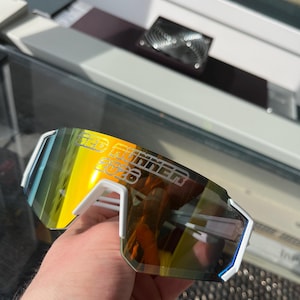 May include: White and blue sports sunglasses with a mirrored yellow and orange lens. The text "RED RUNNER 2026" is printed on the lens. The sunglasses have a modern, angular design.