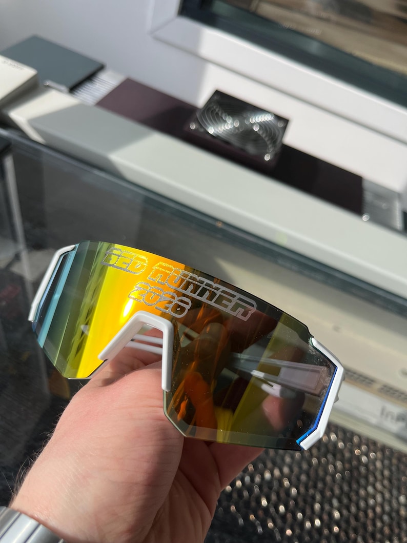 May include: White and blue sports sunglasses with a mirrored yellow and orange lens. The text "RED RUNNER 2026" is printed on the lens. The sunglasses have a modern, angular design.