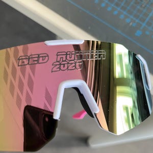 May include: White and pink sports sunglasses with mirrored lenses. The text "RED RUNNER 2020" is printed on the lens. The sunglasses have a white frame and a pink nose piece.