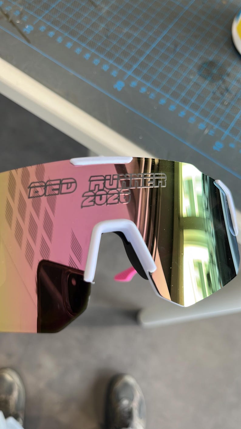 May include: White and pink sports sunglasses with mirrored lenses. The text "RED RUNNER 2020" is printed on the lens. The sunglasses have a white frame and a pink nose piece.