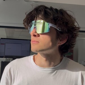 May include: White-framed sunglasses with a mirrored, multi-colored lens. The sunglasses have the word "FACTORY" printed on the lens. The person is wearing a white t-shirt.