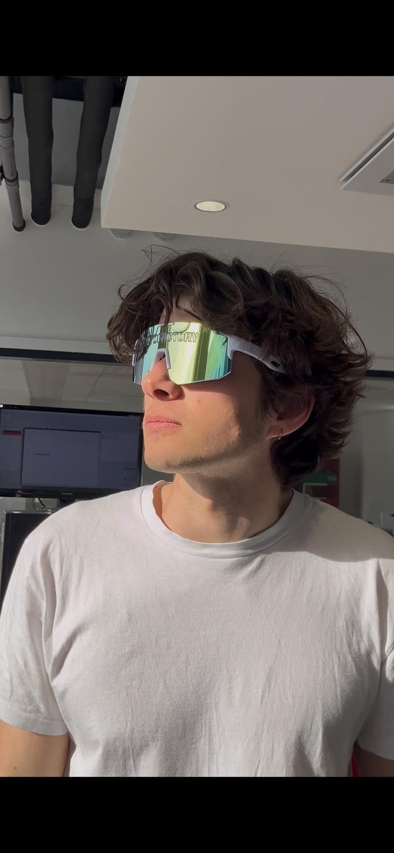 May include: White-framed sunglasses with a mirrored, multi-colored lens. The sunglasses have the word "FACTORY" printed on the lens. The person is wearing a white t-shirt.