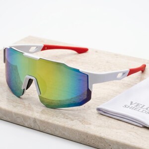 May include: White sports sunglasses with a mirrored, multi-colored lens. The arms are white with red tips. A white cloth case with the text "VELO SHIELDS" is next to the sunglasses.