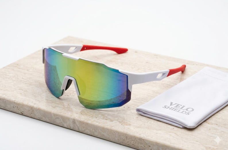 May include: White sports sunglasses with a mirrored, multi-colored lens. The arms are white with red tips. A white cloth case with the text "VELO SHIELDS" is next to the sunglasses.